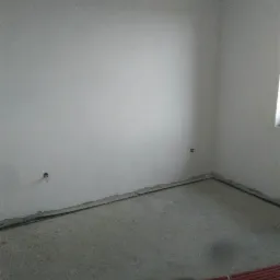 Surowe wnętrze pokoju w trakcie remontu: ściany pokryte świeżym tynkiem, widoczne otwory na gniazdka elektryczne, podłoga betonowa z ułożonymi rurami instalacyjnymi w kolorze czerwonym.