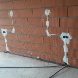 Surowa ściana z cegły z widocznymi, częściowo zamaskowanymi przewodami elektrycznymi i puszkami instalacyjnymi, zabezpieczonymi białą masą uszczelniającą.