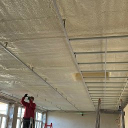 BUD-MAT - Pracownik na drabinie aplikuje pianę izolacyjną na sufit z metalową konstrukcją, widoczny laserowy poziom, okna w tle.