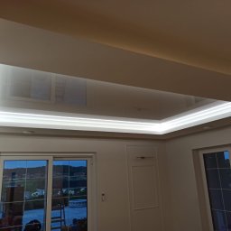 BUD-MAT - Biały, podwieszany sufit z oświetleniem LED w kształcie prostokąta, odbijający światło z okien. Widok z dołu na wnętrze pokoju z oknami i drzwiami balkonowymi.