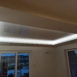 Biały, podwieszany sufit z oświetleniem LED w kształcie prostokąta, odbijający światło z okien. Widok z dołu na wnętrze pokoju z oknami i drzwiami balkonowymi.