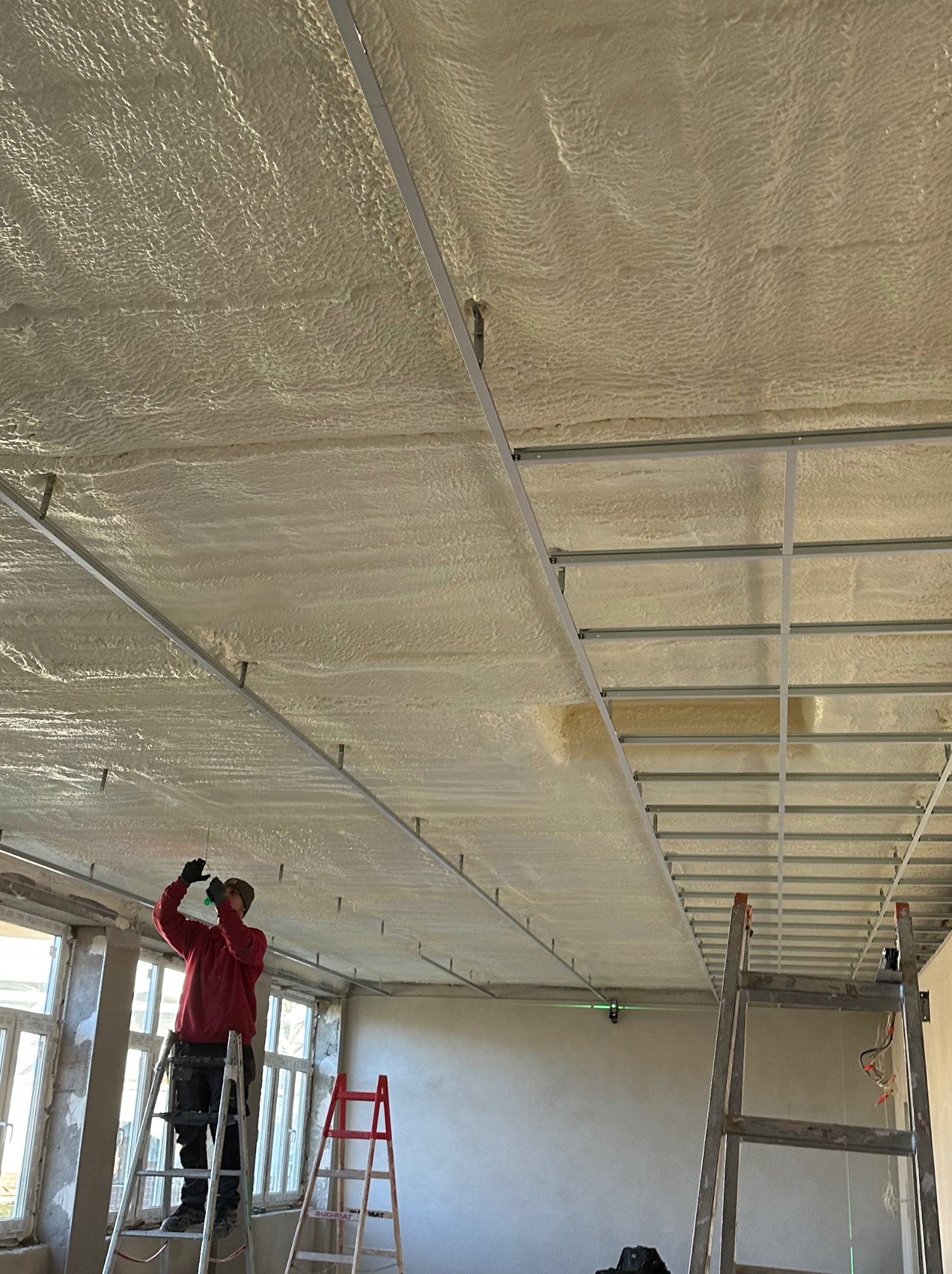 Pracownik na drabinie aplikuje pianę izolacyjną na sufit z metalową konstrukcją, widoczny laserowy poziom, okna w tle.