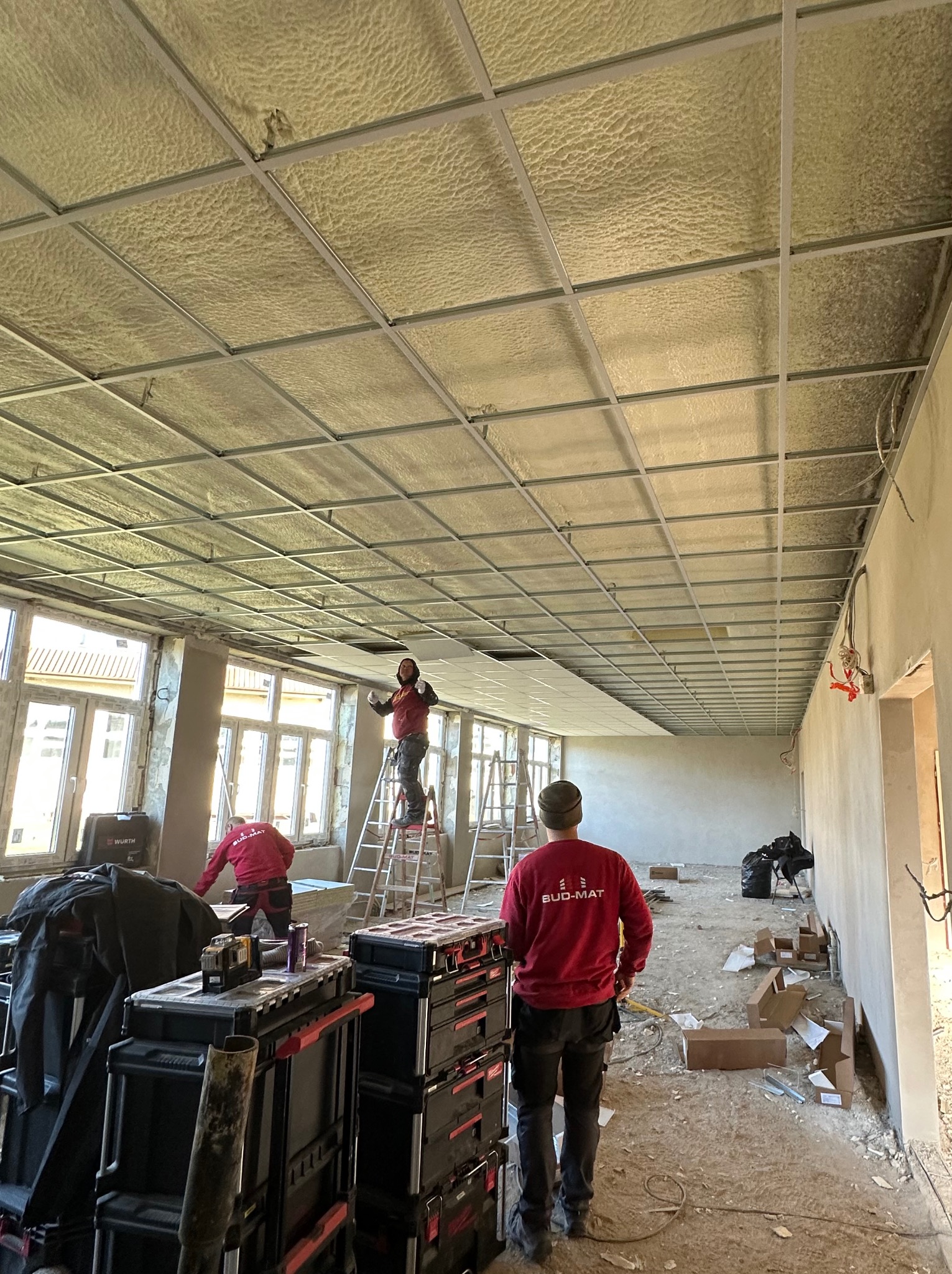 Montaż sufitu podwieszanego z płyt karton-gips w pomieszczeniu z dużymi oknami; pracownicy na drabinie przytwierdzają płyty do metalowej konstrukcji, widoczne narzędzia i skrzynki narzędziowe.