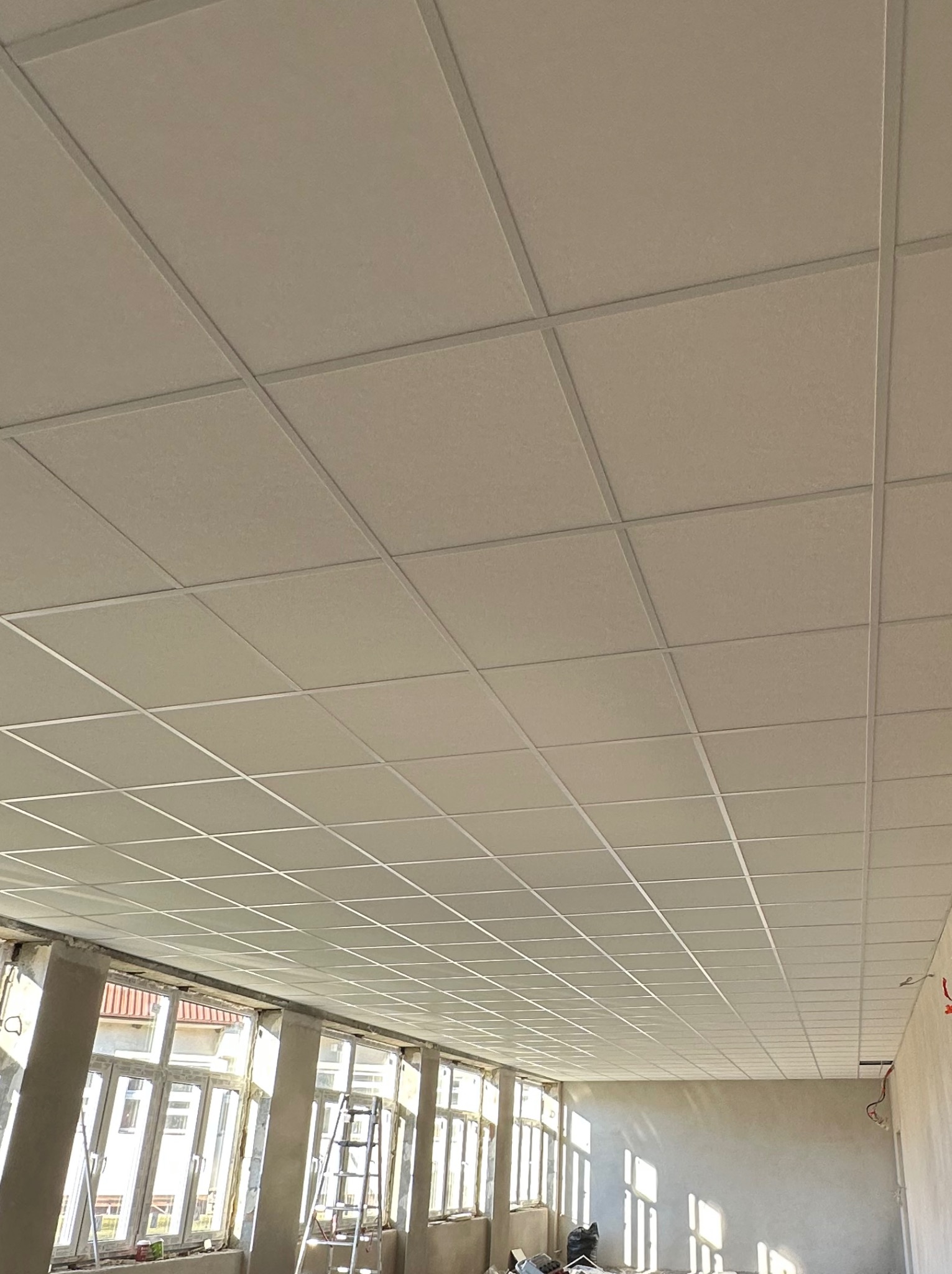 Sufit podwieszany kasetonowy z widoczną konstrukcją metalową, w tle okna i drabina, wnętrze w trakcie wykańczania.