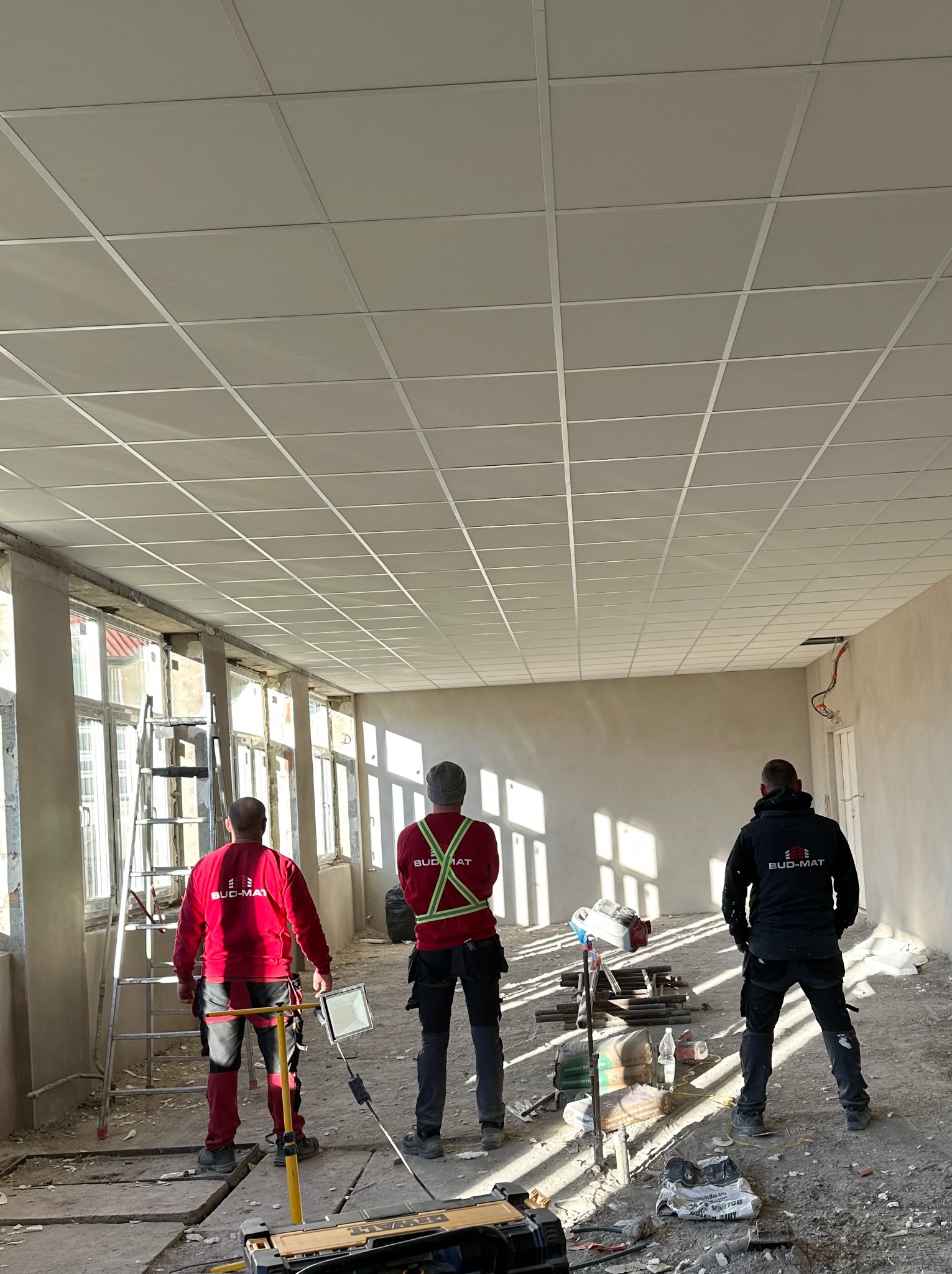 Trzech pracowników firmy Bud-Mat stoi w dużym, jasnym pomieszczeniu z częściowo zamontowanym sufitem podwieszanym, widoczne narzędzia i materiały budowlane, podłoga w trakcie prac wykończeniowych.