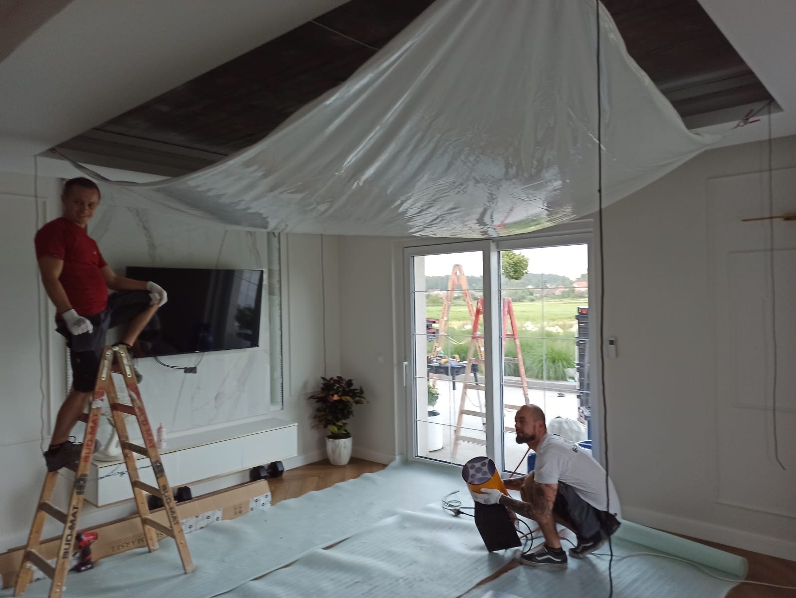Montaż sufitu napinanego w salonie: dwaj monterzy, folia ochronna, drabina Bud-Mat, widok na okno tarasowe i ogród. Prace w toku, zabezpieczone podłoże.