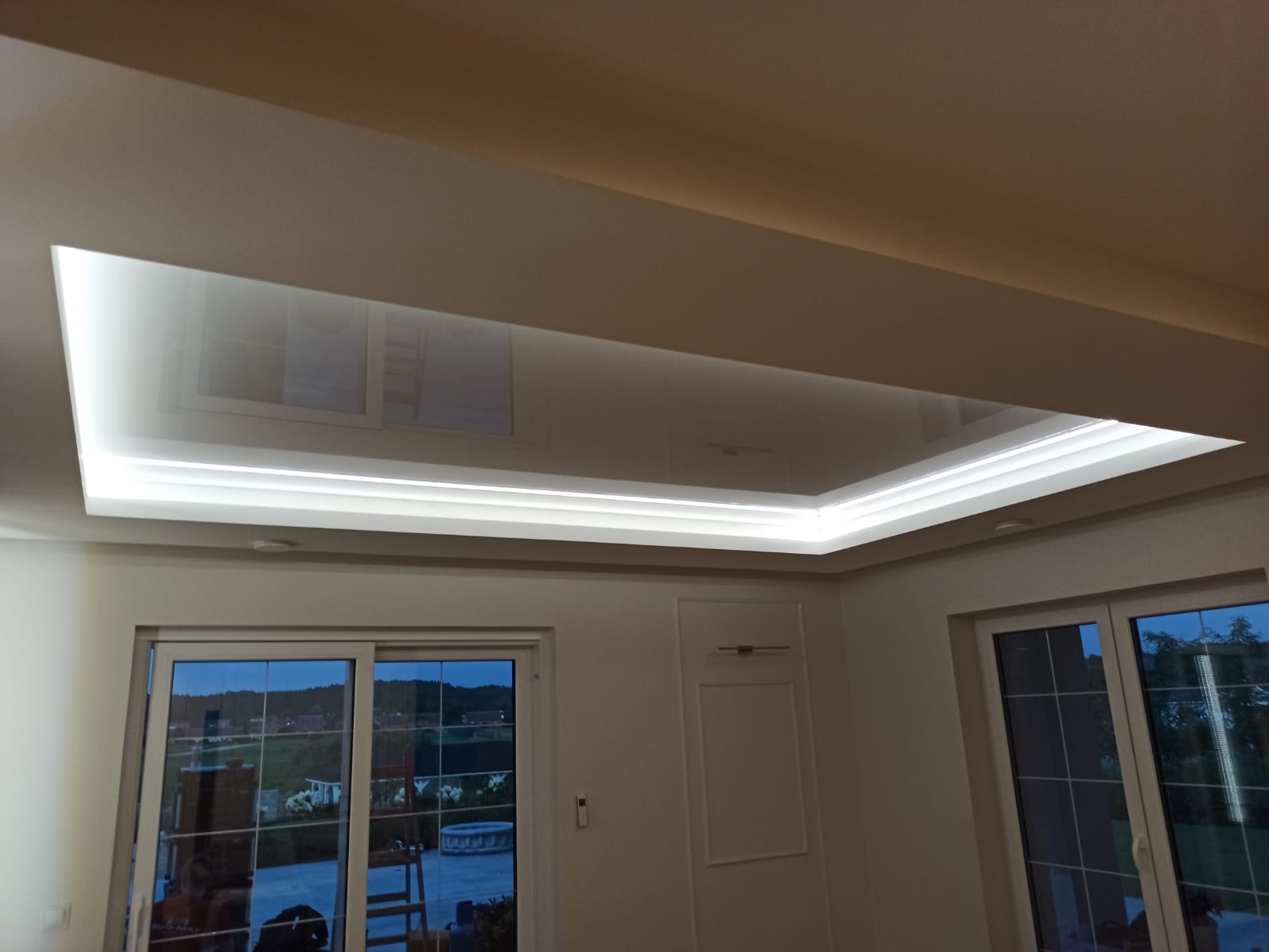 Biały, podwieszany sufit z oświetleniem LED w kształcie prostokąta, odbijający światło z okien. Widok z dołu na wnętrze pokoju z oknami i drzwiami balkonowymi.