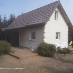 Jasna elewacja domu jednorodzinnego z brązowym dachem i drewnianym tarasem, widoczna z poziomu gruntu, z dekoracyjnymi krzewami i elementami wykończenia.