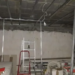 Montaż metalowej konstrukcji sufitu podwieszanego w surowym pomieszczeniu, widoczne profile, drabina i materiały budowlane.