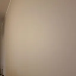 Gładka, beżowa ściana wewnętrzna po malowaniu, widoczna z niskiego kąta, z fragmentem sufitu i mebla w dolnym rogu.