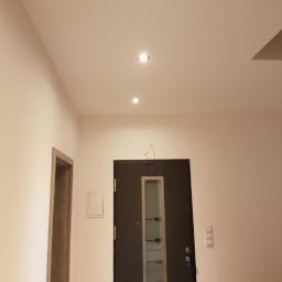 USŁUGI REMONTOWO BUDOWLANE REM - PRUS EWELINA PRUSAK - Wnętrze korytarza po remoncie: świeżo pomalowane ściany w odcieniach bieli, ciemne drzwi wejściowe z przeszkleniami, zamontowane oświetlenie punktowe w suficie, widoczny fragment ościeżnicy.
