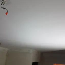 Wnętrze w trakcie prac wykończeniowych, gładko pomalowany sufit z wystającymi przewodami elektrycznymi, ściany częściowo przygotowane pod malowanie z widocznymi śladami szpachlowania.