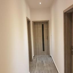 USŁUGI REMONTOWO BUDOWLANE REM - PRUS EWELINA PRUSAK - Wyremontowany korytarz z szarymi panelami podłogowymi imitującymi drewno, kremowymi ścianami i dwiema drewnianymi ościeżnicami drzwi, oświetlony trzema punktowymi lampami sufitowymi.