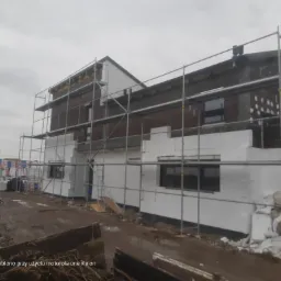 Budowa domu w Szczepanowie w trakcie ocieplania elewacji, widoczne rusztowania i styropian na ścianach, teren wokół budynku błotnisty.