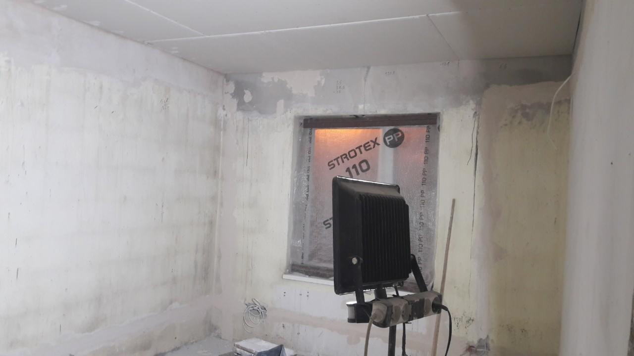 Pokój w trakcie remontu: ściany z częściowo nałożoną gładzią, okno zabezpieczone folią Strotex PP 110, przenośny reflektor oświetlający wnętrze.