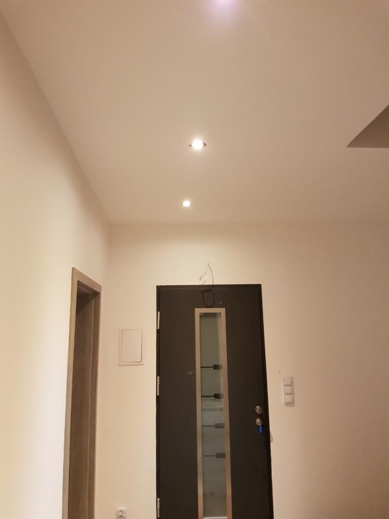 Wnętrze korytarza po remoncie: świeżo pomalowane ściany w odcieniach bieli, ciemne drzwi wejściowe z przeszkleniami, zamontowane oświetlenie punktowe w suficie, widoczny fragment ościeżnicy.