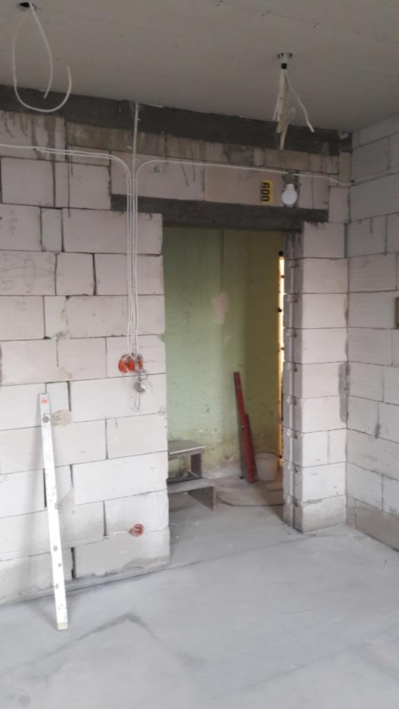 Surowy stan wnętrza z białymi bloczkami, widoczne instalacje elektryczne, poziomica oparta o ścianę i surowy beton na podłodze.