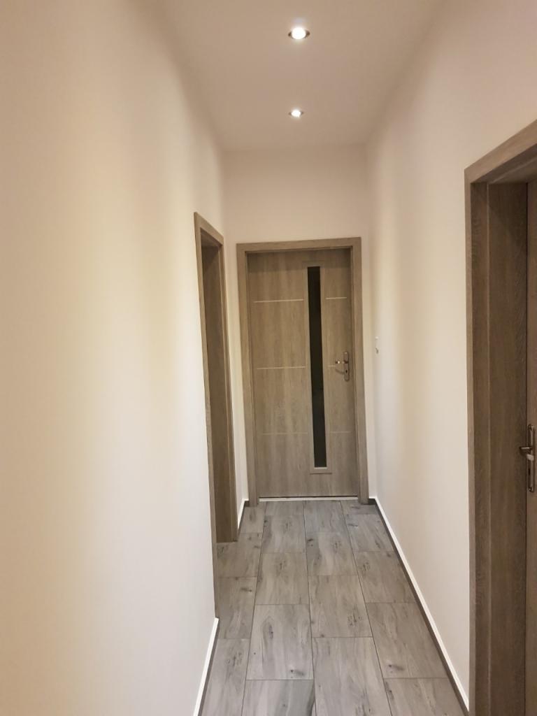 Wyremontowany korytarz z szarymi panelami podłogowymi imitującymi drewno, kremowymi ścianami i dwiema drewnianymi ościeżnicami drzwi, oświetlony trzema punktowymi lampami sufitowymi.