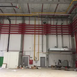 Wnętrze hali przemysłowej z rozbudowaną siecią rur w kolorach czerwonym i żółtym pod sufitem, widoczny system przeciwpożarowy, wentylacja i instalacje elektryczne, na podłodze resztki materiałów...