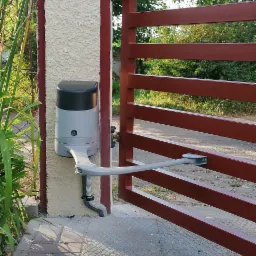 Ciemnoczerwona brama uchylna z automatyką, zamontowana na słupku z jasnym tynkiem strukturalnym i czerwoną obwódką, widok z bliska na mechanizm otwierający.