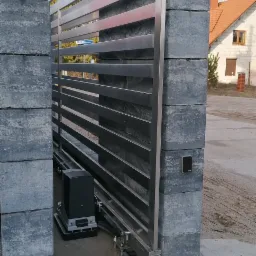 Nowoczesna brama przesuwna z poziomych profili, osadzona na murowanych słupkach z szarego kamienia, widoczny napęd i elementy automatyki, w tle dom jednorodzinny.
