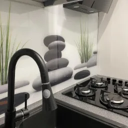 Czarny, granitowy zlew kuchenny z baterią, na tle szklanego panelu z motywem kamieni i trawy, obok płyta gazowa.