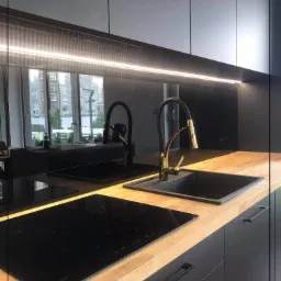 Nowoczesna kuchnia z czarnymi szafkami, drewnianym blatem, złotą baterią i szklanym panelem nad blatem, odbijającym okno.