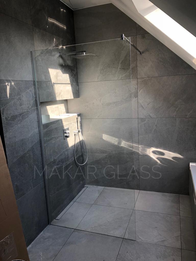 Nowoczesna kabina prysznicowa ze szklaną ścianą i deszczownicą, zamontowana w łazience z szarymi płytkami imitującymi kamień, oświetlona naturalnym światłem wpadającym przez okno dachowe.