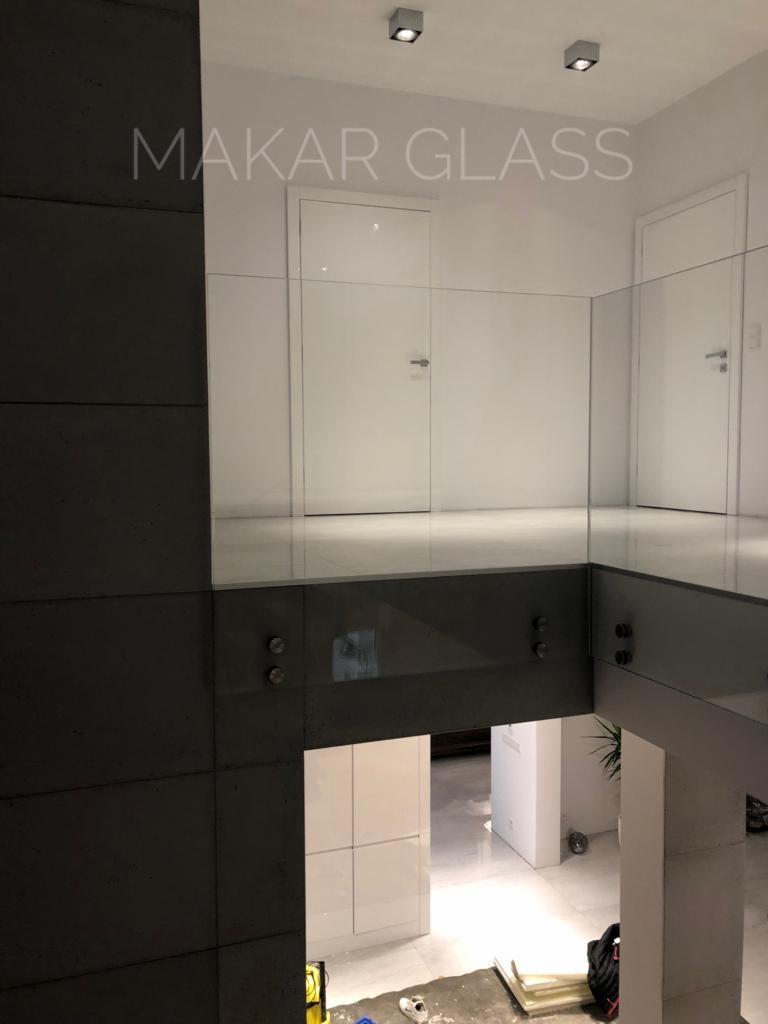 Szklana balustrada z metalowymi mocowaniami, widok z góry, widoczne dwie białe drzwi w tle, logo firmy Makar Glass na szkle.