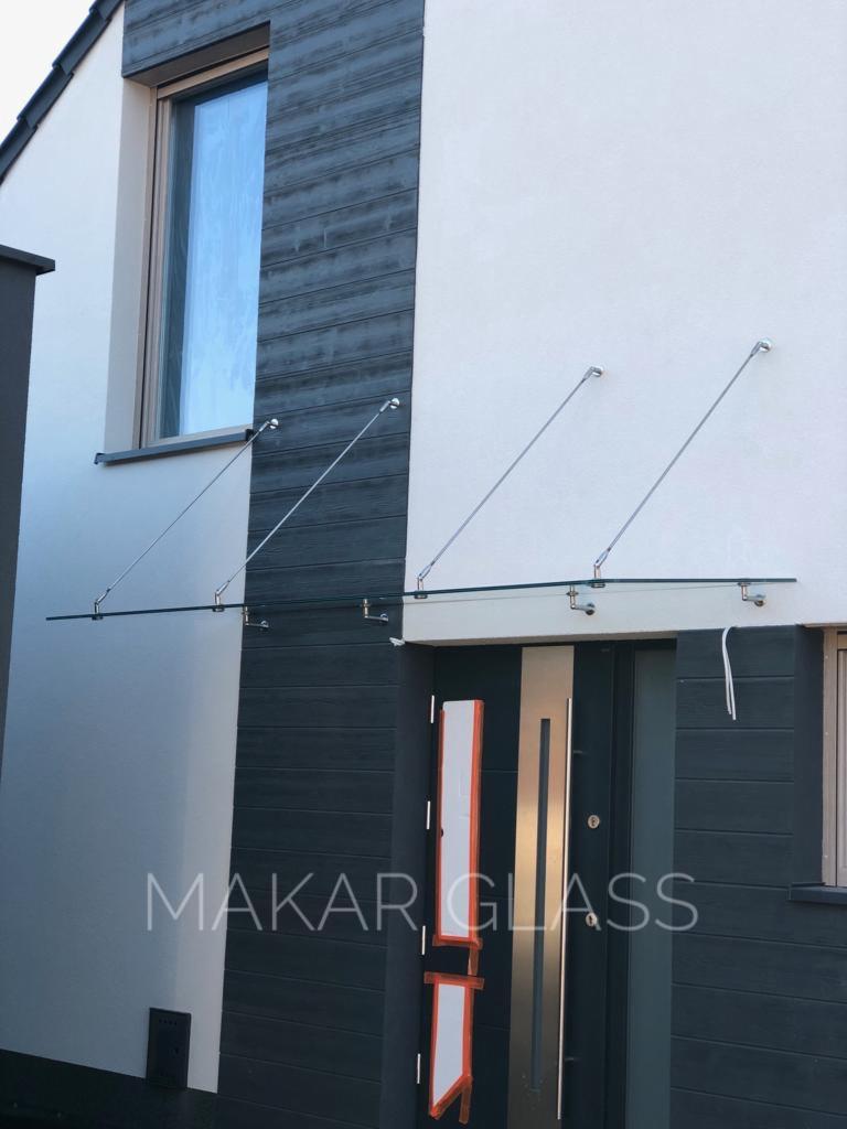 Szklane zadaszenie nad nowoczesnymi drzwiami wejściowymi, wsparte na metalowych prętach, widoczne logo 'MAKAR GLASS' na elewacji.