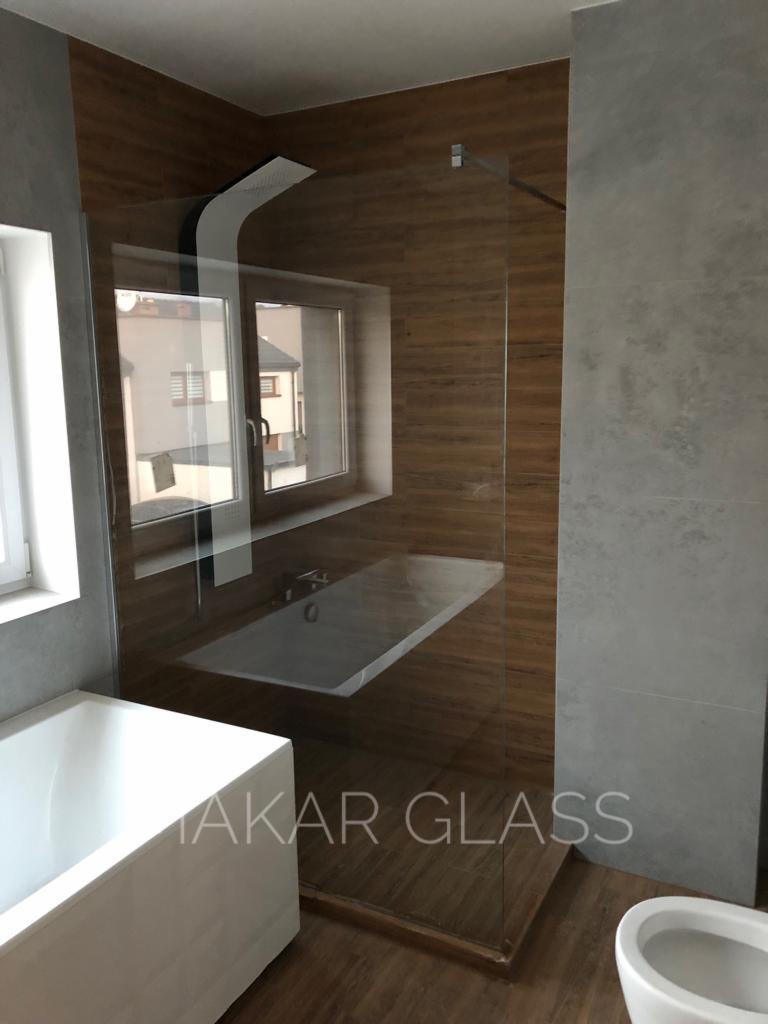 Nowoczesna kabina prysznicowa ze szkła hartowanego, z deszczownicą i widoczną wanną obok, na ścianie brązowe płytki imitujące drewno, na szklanej ściance widoczne logo firmy.