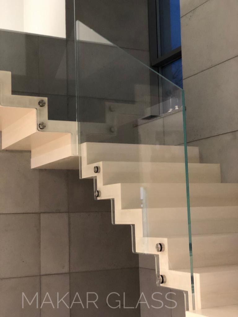 Nowoczesne schody z jasnego drewna z przezroczystą balustradą szklaną, mocowaną metalowymi śrubami, na tle ściany z szarych płyt, widoczne logo 'MAKAR GLASS'.