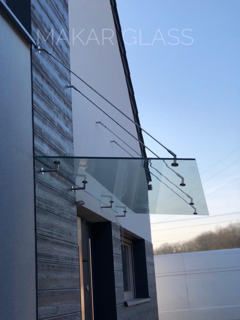 Szklany daszek nad wejściem do domu, mocowany na metalowych wspornikach, z widocznym logo 'MAKAR GLASS' w górnej części zdjęcia.