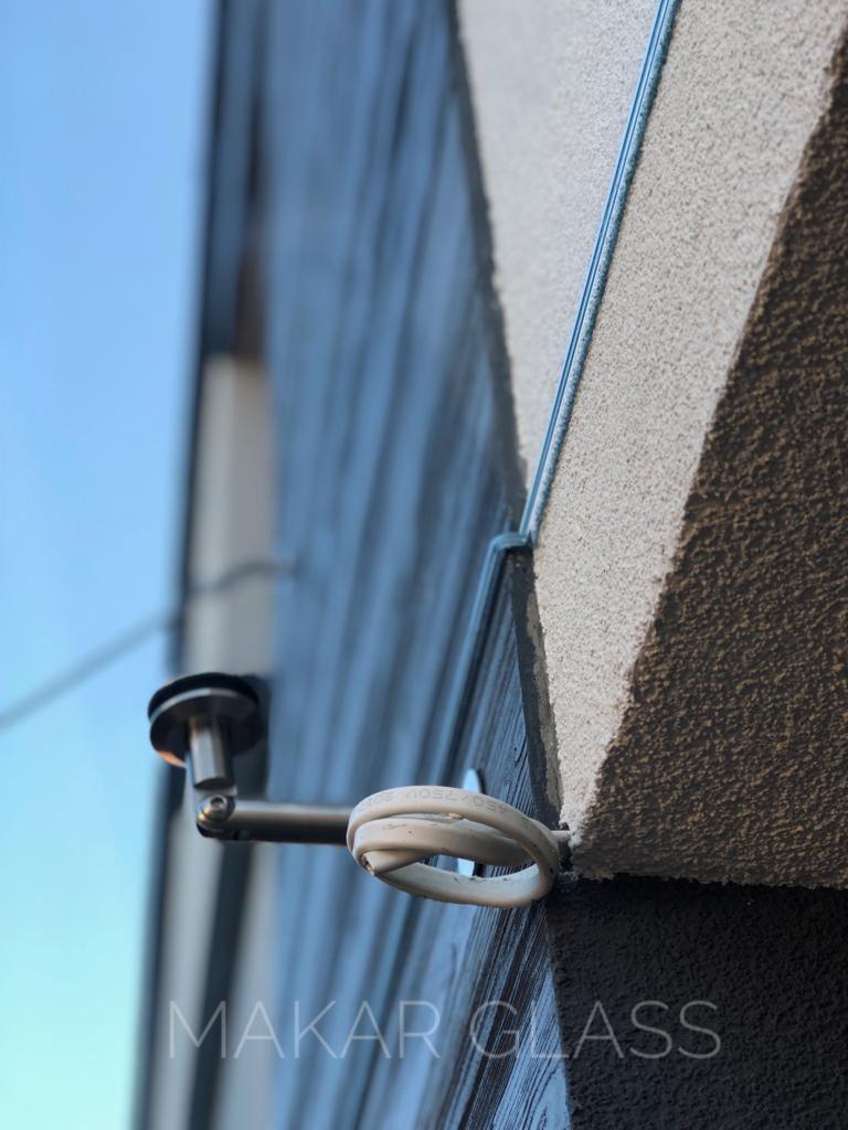 Zewnętrzny narożnik budynku z zamontowaną szklaną płytą, oświetloną lampą na metalowym ramieniu i widocznym okablowaniem.