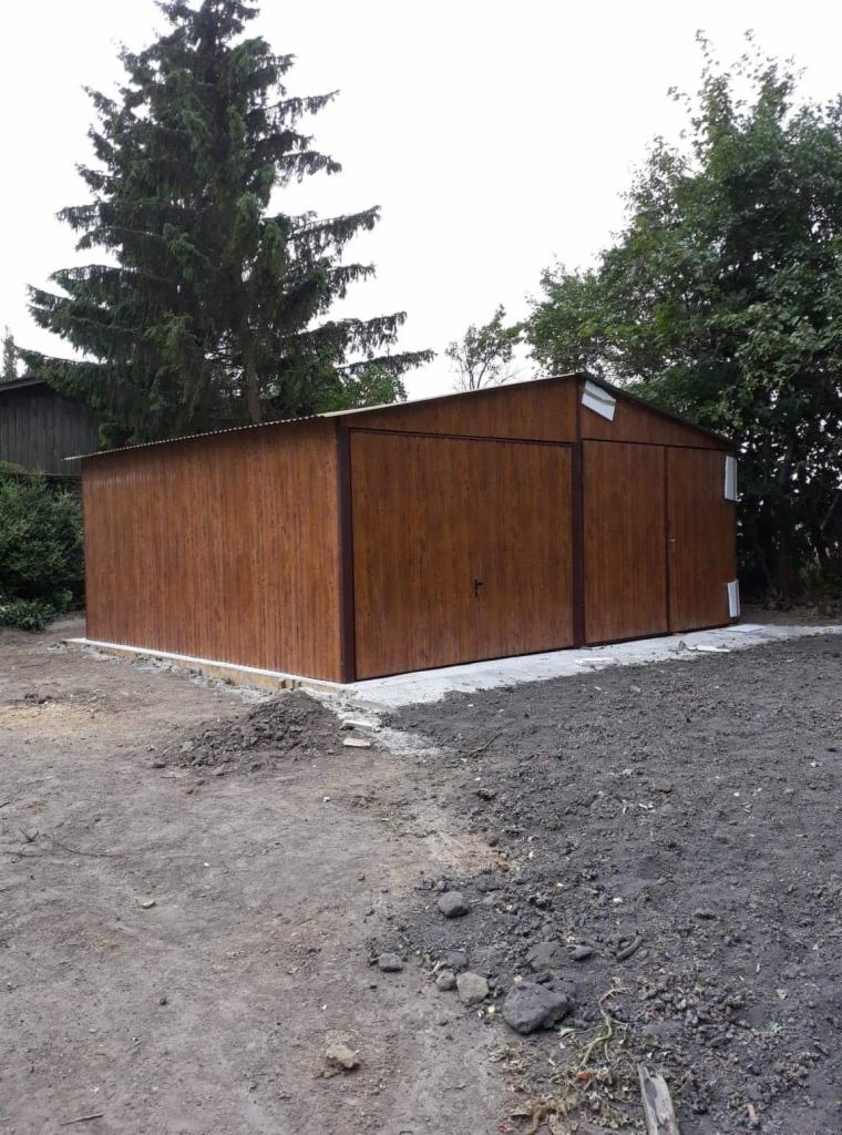 Brązowy garaż blaszany z dwoma bramami i drzwiami, osadzony na betonowej wylewce, na tle drzew i pochmurnego nieba.
