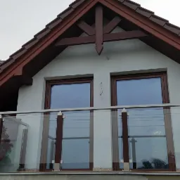 Szklana balustrada balkonowa z brązowymi słupkami i stalowymi elementami mocującymi, widok z dołu na tle dwuspadowego dachu z brązową dachówką i drewnianym ozdobnym szczytem.