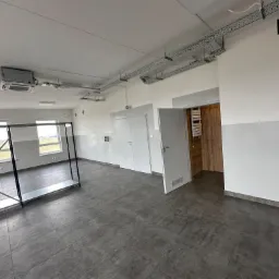 Wnętrze pomieszczenia po remoncie z szarą podłogą z płytek, świeżo pomalowanymi ścianami z szarym pasem na dole, widocznymi instalacjami pod sufitem oraz ramą metalową w lewej części kadru.