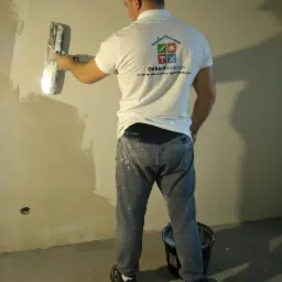 Mężczyzna w białej koszulce z logo firmy Deluxehome.pl, w szarych spodniach z plamami farby, szpachluje ścianę metalową pacą, obok stoi czarne wiadro z materiałem.