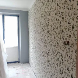 Ściana pokoju pokryta tapetą w motywy roślinne, z widocznym gniazdkiem elektrycznym i fragmentem przygotowanego do malowania okna oraz zabezpieczoną podłogą.