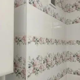 Białe, błyszczące płytki ścienne z motywem kwiatowym w łazience, widoczny fragment wentylacji i szafki.