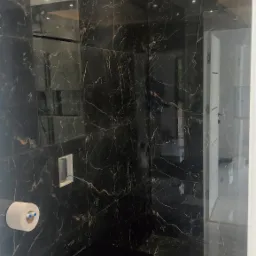 Wykończona łazienka z czarnymi płytkami imitującymi marmur ze złotymi żyłami, podwieszana toaleta w czarnym kolorze, biała podłoga, oświetlenie sufitowe LED i lustro na ścianie.