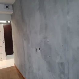 Ściana w szarym odcieniu z efektem dekoracyjnym, widoczne puszki elektryczne bez osprzętu, fragment podłogi z drewnianą listwą przypodłogową oraz drzwi w tle.