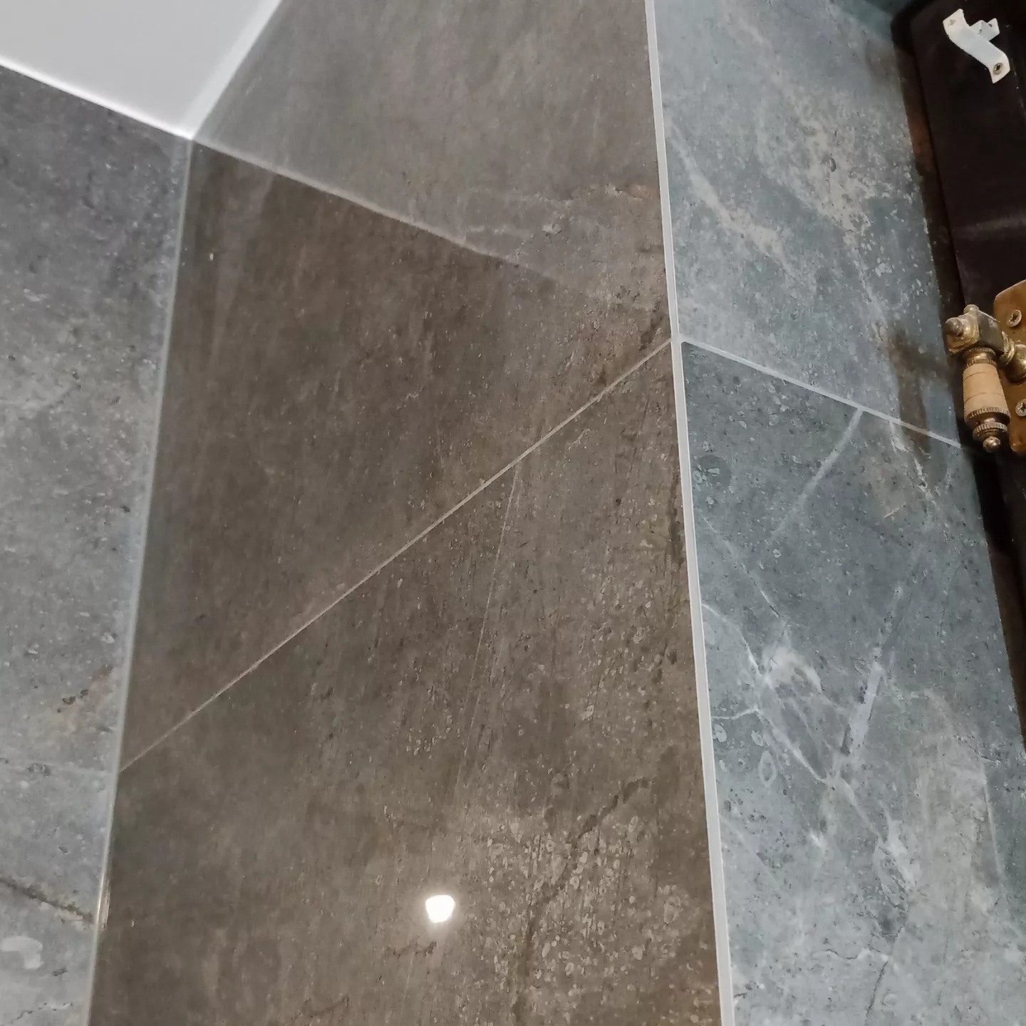 Fragment ściany wyłożonej szarymi płytkami imitującymi marmur, widoczna fuga i zawias drzwi w prawym górnym rogu.