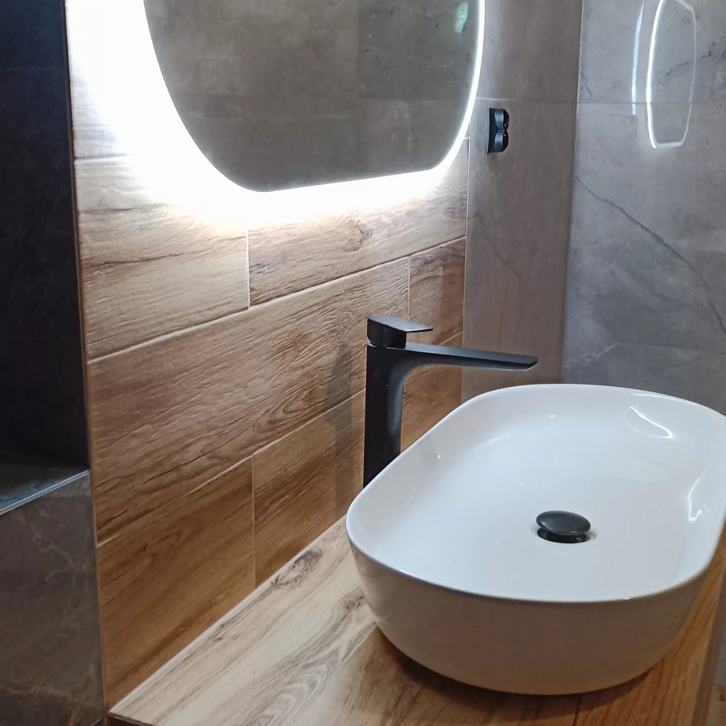Nowoczesna umywalka nablatowa w łazience z podświetlanym lustrem i okładziną imitującą drewno na ścianie.