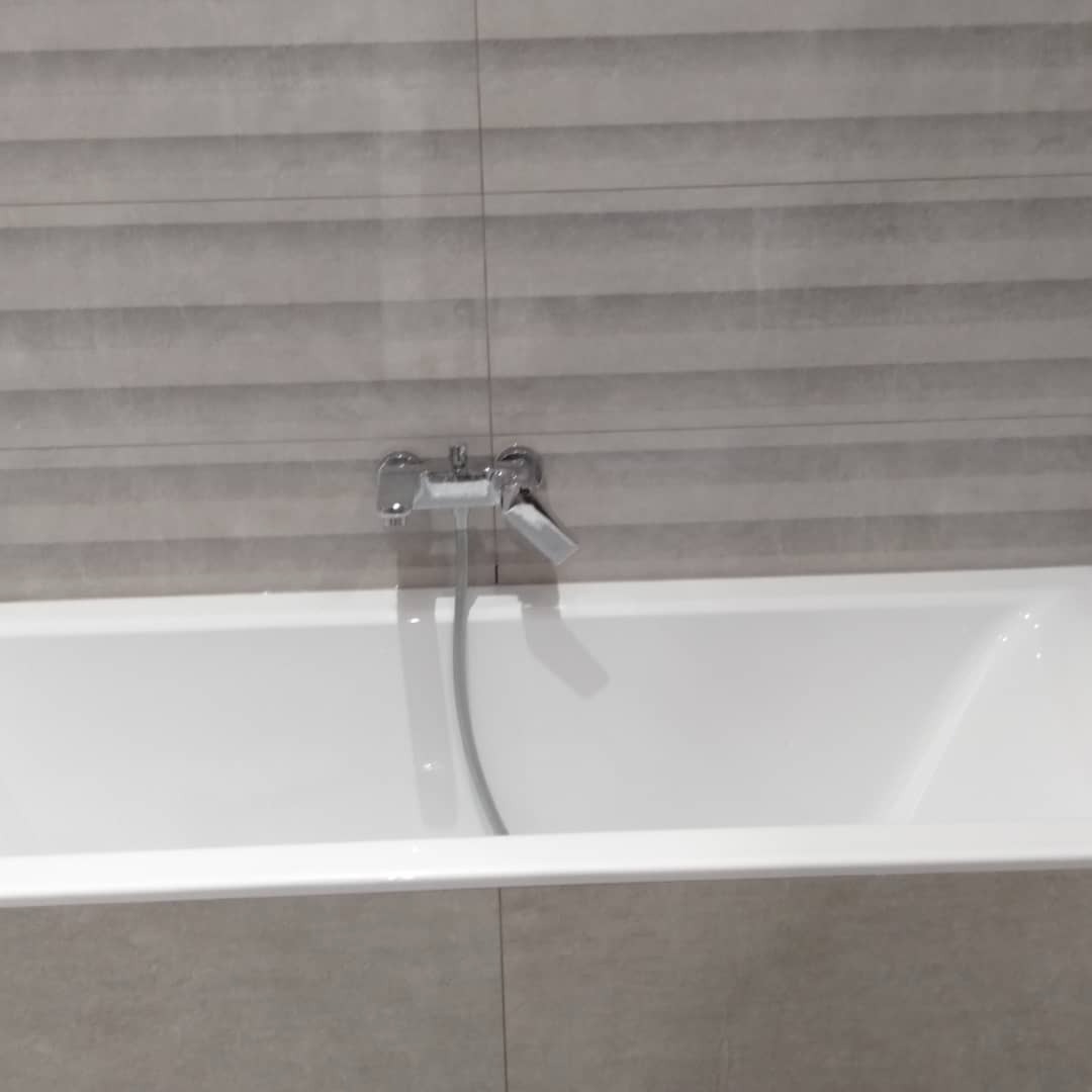 Biała wanna z chromowaną baterią na tle szarych, poziomych płytek imitujących kamień z delikatnym wzorem.