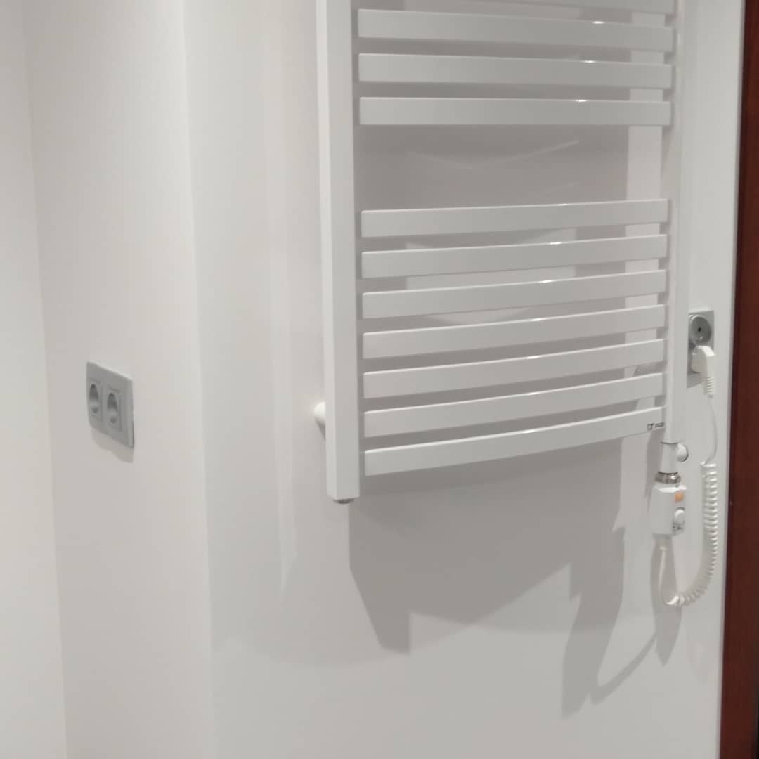 Biały, elektryczny grzejnik drabinkowy zamontowany na białej ścianie w łazience, widoczne gniazdko elektryczne i regulator temperatury.