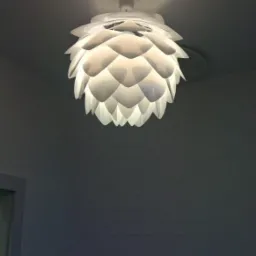 Biała, nowoczesna lampa sufitowa w kształcie szyszki, oświetlająca wzorzystą ścianę w odcieniach szarości, z widocznym czujnikiem dymu na suficie.