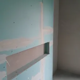 Wnętrze w trakcie prac wykończeniowych: ściana z zamontowaną płytą gipsowo-kartonową z wyciętą wnęką, widoczne szpachlowanie łączeń, surowa podłoga.