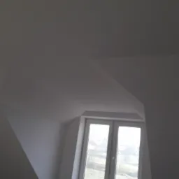 Poddasze po remoncie z oknem dachowym, białe ściany i sufit, widok na zewnątrz przez okno.