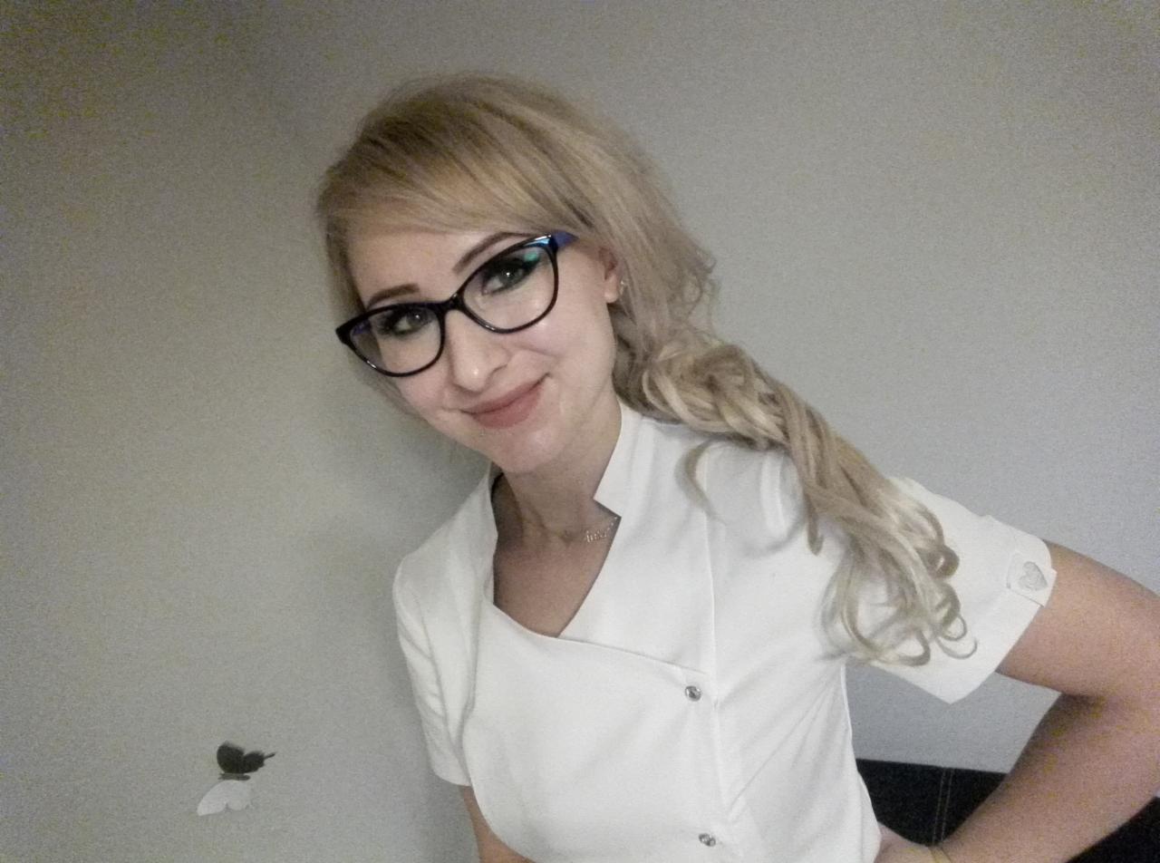 Uśmiechnięta blondynka w okularach i białym uniformie z krótkim rękawem, pozująca na tle szarej ściany z naklejką motyla.